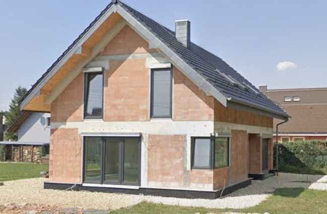 Dom w trakcie budowy z zamontowanymi oknami i drzwiami przesuwnymi. Widoczne bloczki ceramiczne, szara dachówka i częściowo zagospodarowany teren.
