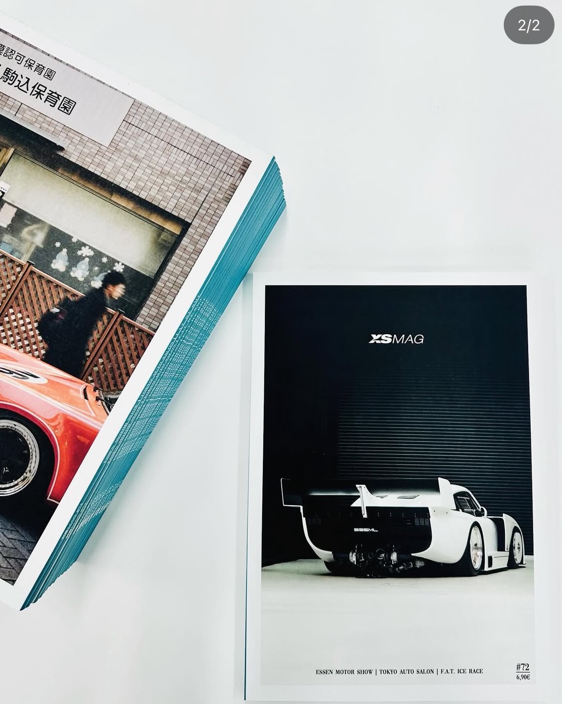 Stos magazynów motoryzacyjnych z okładką czerwonego auta i pojedynczy egzemplarz z białym samochodem wyścigowym na tle czarnej ściany z napisem XSMAG.