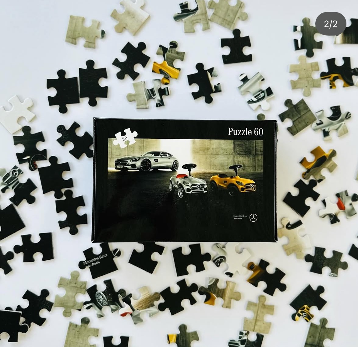 Pudełko puzzli 60-elementowych z motywem Mercedes-Benz, otoczone rozrzuconymi elementami układanki na białym tle. Widoczne fragmenty samochodów na puzzlach.
