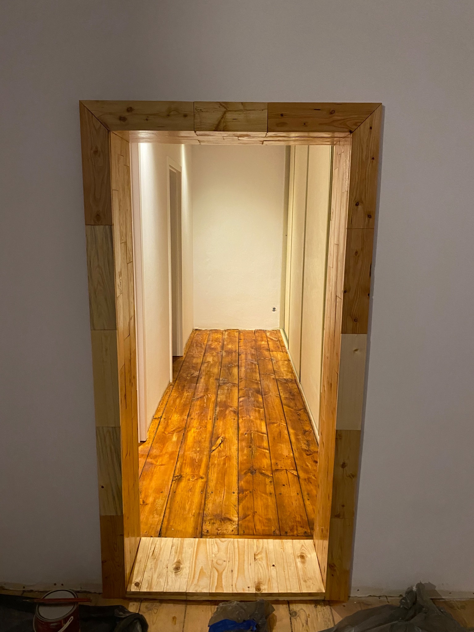 Drewniana futryna z surowych desek i odnowiona podłoga z desek w długim korytarzu. Widoczne ślady prac remontowych i wiaderko z farbą.