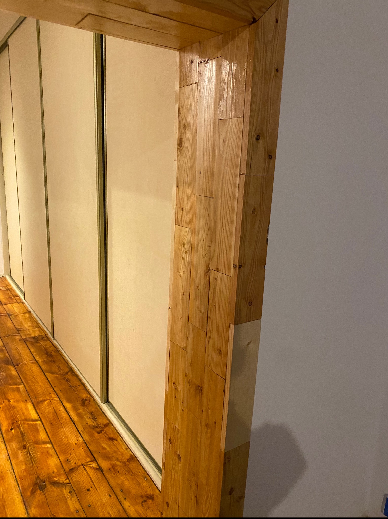 Wykończenie drewnianej futryny w pokoju z przesuwną szafą, widoczna podłoga z desek i fragment ściany. Elementy drewniane lakierowane.