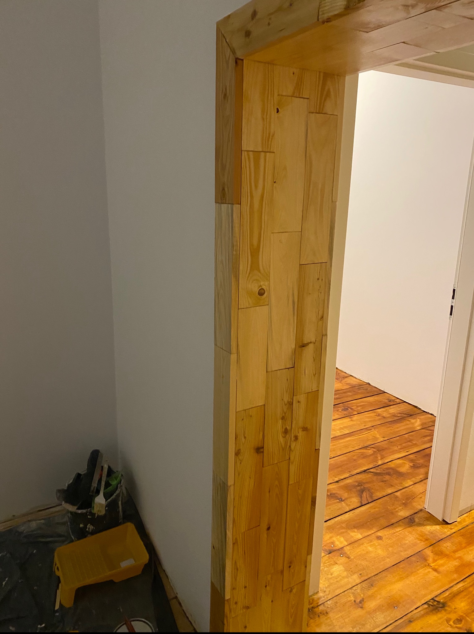 Wykończone drewnem obramowanie drzwi z widocznymi narzędziami malarskimi w rogu, ukazujące etap prac podczas wykańczania wnętrza z drewnianą podłogą.