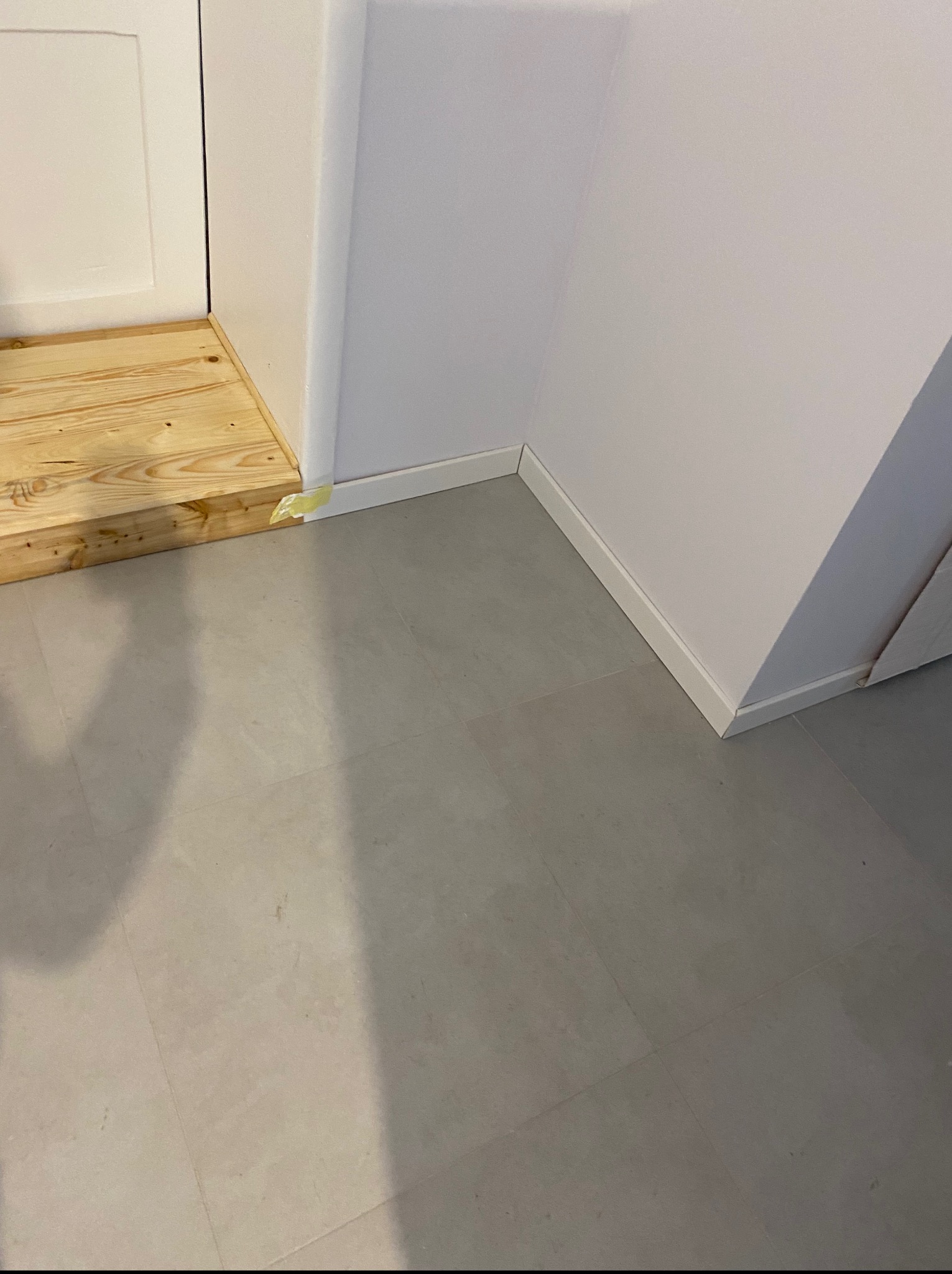 Szare płytki podłogowe, białe listwy przypodłogowe i drewniany próg przy białych drzwiach. Widoczny fragment taśmy malarskiej.