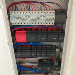 Wnętrze białej szafy rozdzielczej z zainstalowanymi modułami, bezpiecznikami i przewodami w kolorach czerwonym, niebieskim i zielonym. Widoczne elementy systemu inteligentnego domu.