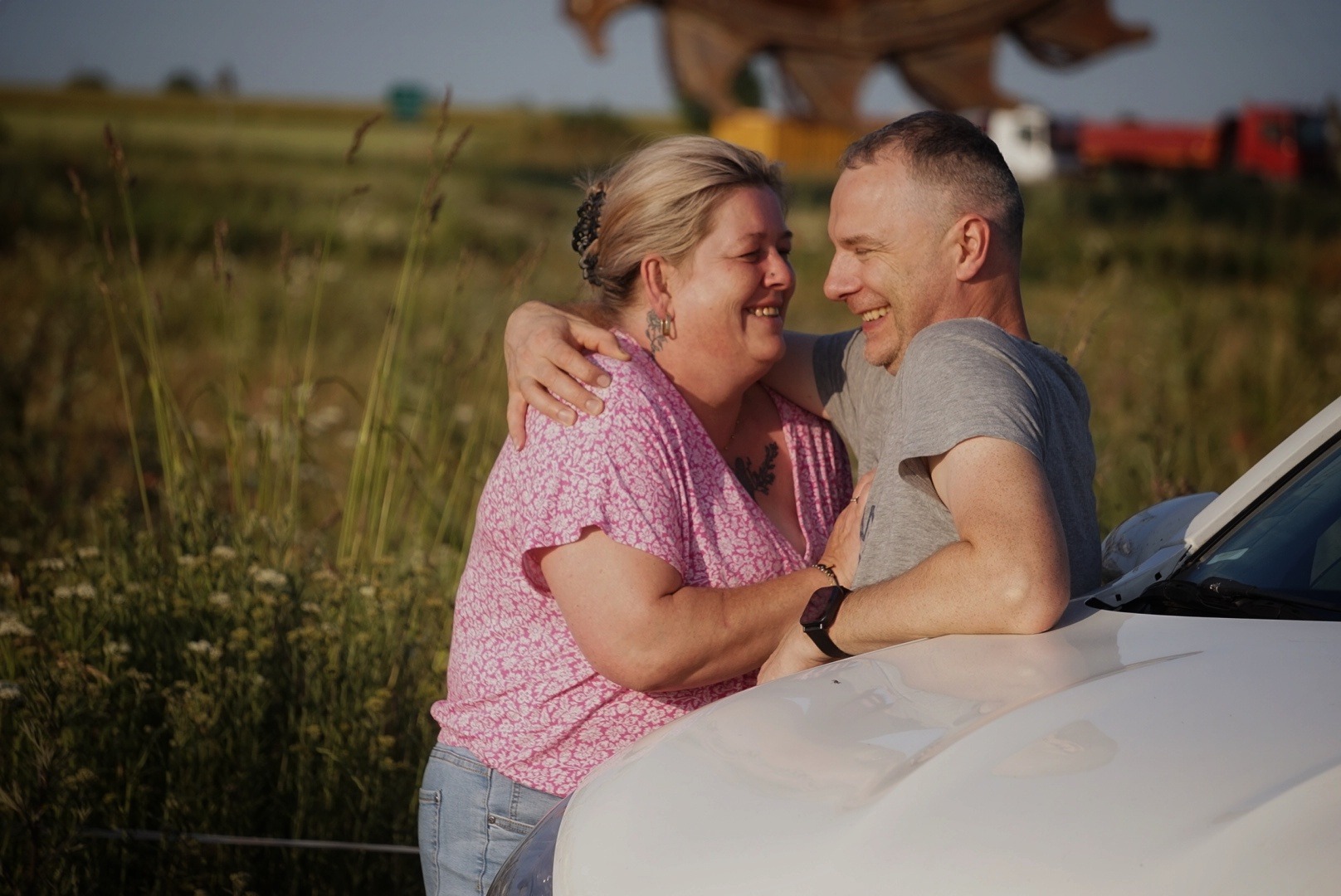 Para uśmiechnięta, obejmująca się przy białym samochodzie na tle łąki i nieba. Kobieta w różowej bluzce, mężczyzna w szarej koszulce. Widoczne tatuaże i biżuteria.