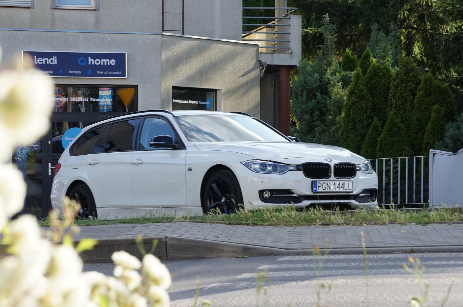 Białe BMW kombi zaparkowane przed budynkiem z szyldem 'lendi home', z widocznymi kwiatami na pierwszym planie i zielenią w tle. Ujęcie z niskiego kąta.