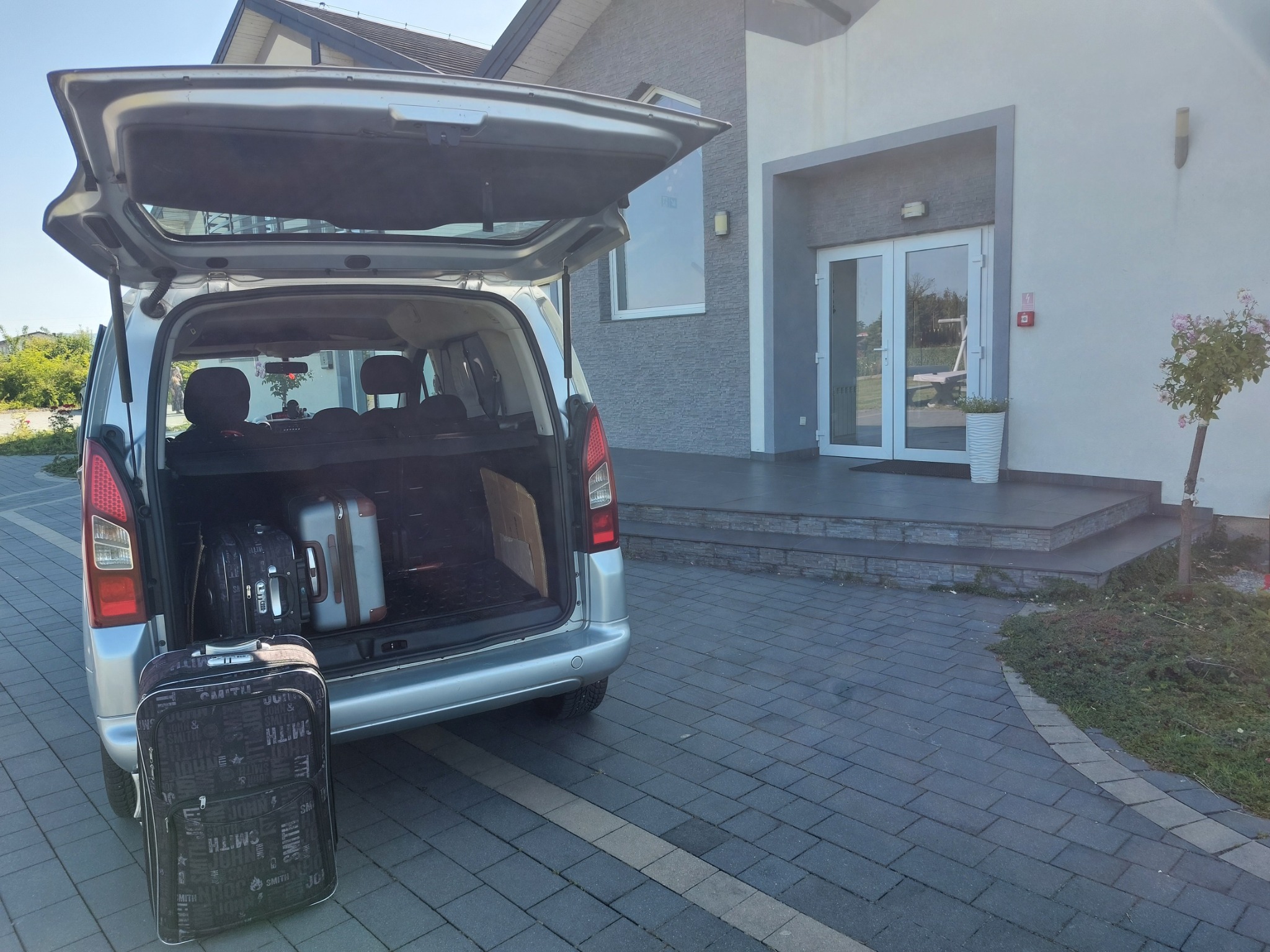 Srebrny van z otwartym bagażnikiem, w którym znajdują się walizki. Przed autem stoi walizka na kółkach. W tle budynek z białymi drzwiami.