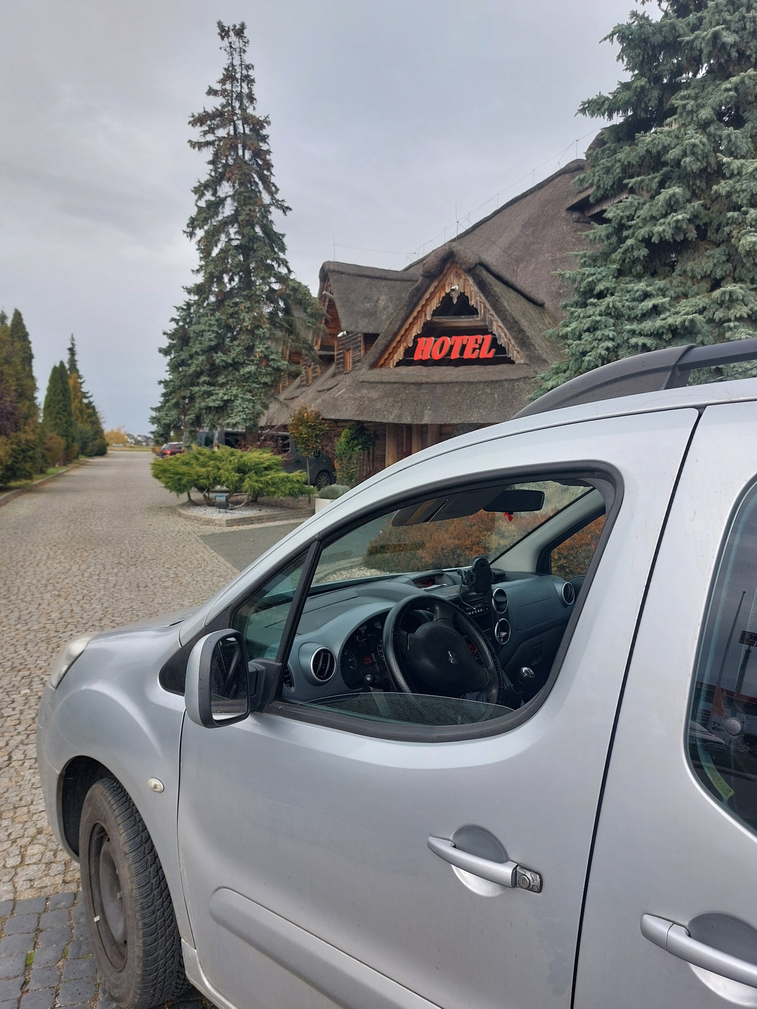 Srebrny samochód osobowy na tle hotelu z strzechą w Żywcu. Widok z boku, częściowo widoczne wnętrze pojazdu. Usługi transportowe dla gości hotelowych.