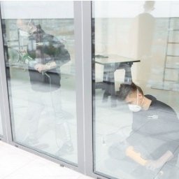 PolerkaSzyb.pl - Dwóch monterów w maskach przy dużym oknie aluminiowym podczas prac wykończeniowych. Jeden szlifuje, drugi klęczy z narzędziami.