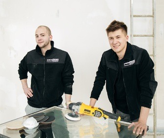 Dwóch mężczyzn w firmowych bluzach poleruje szybę szlifierką kątową na tle drabiny i ściany w trakcie prac wykończeniowych.