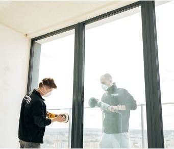 Pracownik w masce szlifuje okno balkonowe z czarną ramą w jasnym wnętrzu, drugi pracownik obok. Widok na miasto za oknem. Praca w toku.