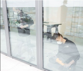 Dwóch monterów w maskach przy dużym oknie aluminiowym podczas prac wykończeniowych. Jeden szlifuje, drugi klęczy z narzędziami.