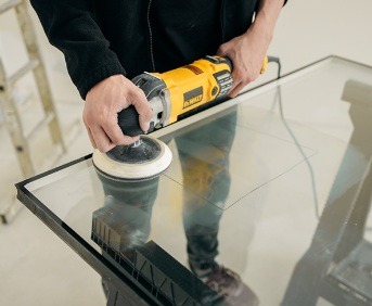 Szlifowanie szyby okiennej szlifierką DeWalt na stole warsztatowym. Widoczna drabina w tle. Precyzyjna obróbka szkła w jasnym pomieszczeniu.