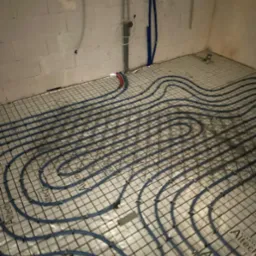 Niebieskie rury ogrzewania podłogowego ułożone na siatce w surowym pomieszczeniu, widoczne pionowe rury instalacji wodnej i elektrycznej przy ścianie.