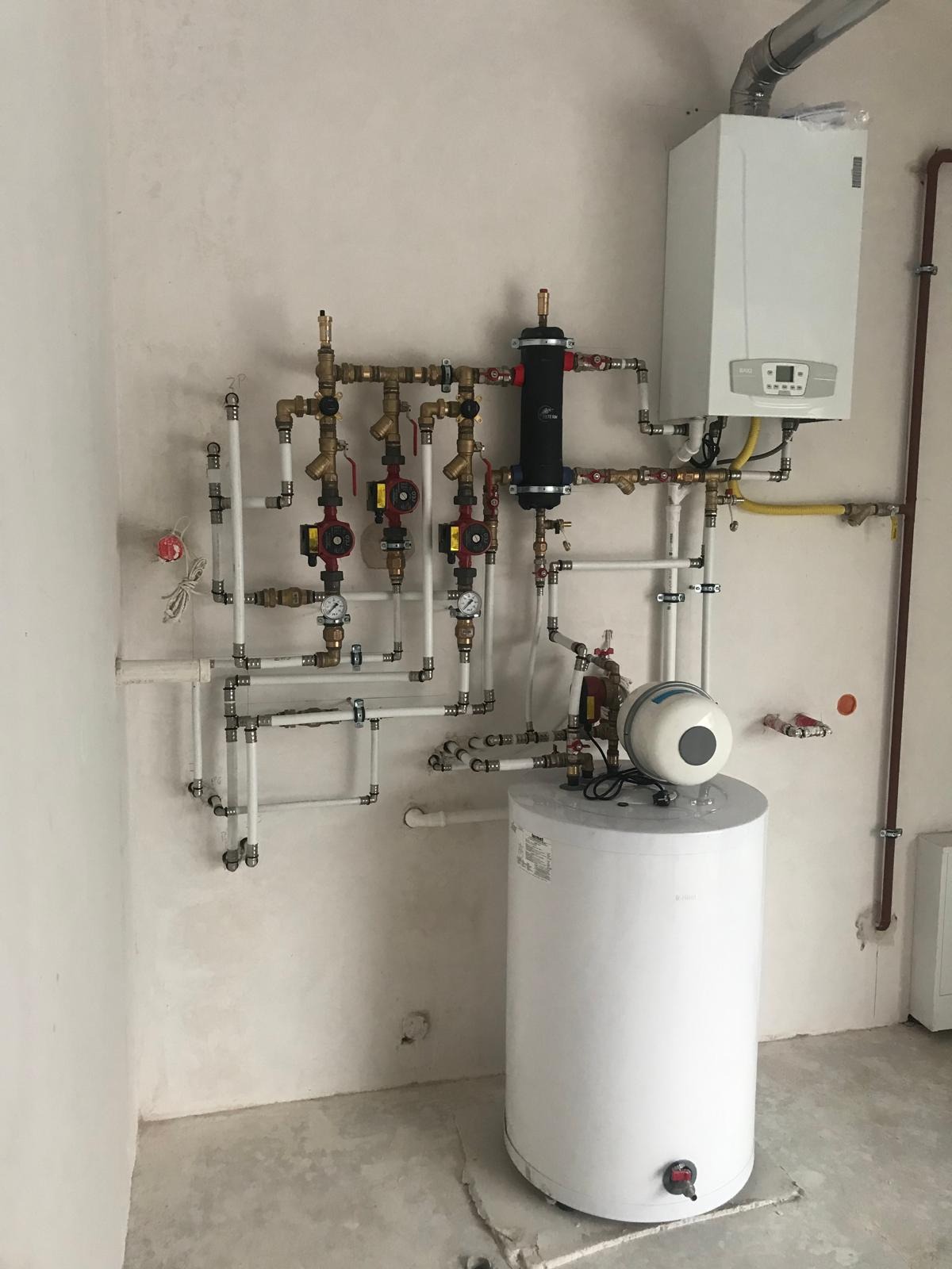 Złożona instalacja grzewcza z kotłem, zbiornikiem i licznymi rurami, zaworami i pompami na ścianie w surowym pomieszczeniu. Widoczne elementy instalacji w kolorze białym i miedzianym.