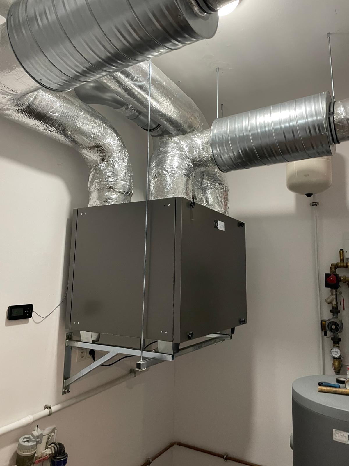 Szara centrala wentylacyjna z podwieszonymi, izolowanymi przewodami wentylacyjnymi w pomieszczeniu technicznym. Widoczny sterownik na ścianie i fragment instalacji hydraulicznej.