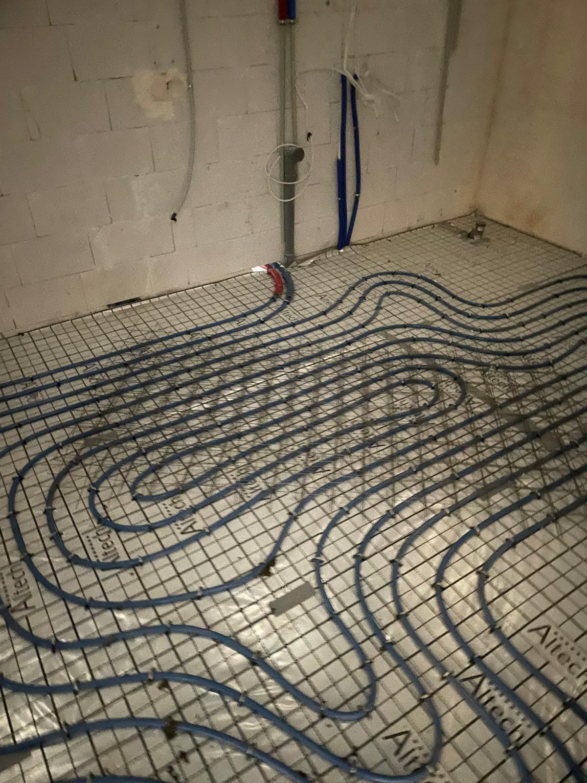 Niebieskie rury ogrzewania podłogowego ułożone na siatce w surowym pomieszczeniu, widoczne pionowe rury instalacji wodnej i elektrycznej przy ścianie.