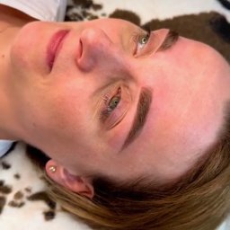 KAROLINA GÓRALEWSKA - Kobieta po zabiegu microbladingu brwi, leżąca na wzorzystym podłożu. Widoczne świeżo wykonane, ciemne brwi i delikatny rumień na skórze.