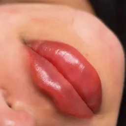 Makro zbliżenie na usta po zabiegu makijażu permanentnego, ukazujące intensywny kolor i połysk. Widoczne delikatne detale skóry wokół ust. Ciemne tło.