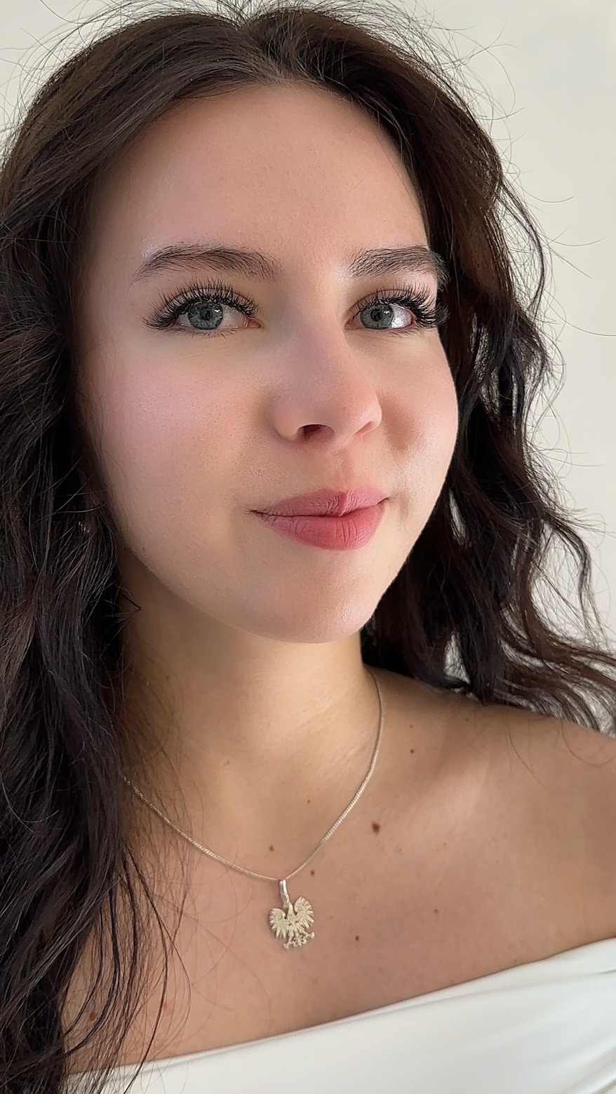Portret kobiety z przedłużonymi rzęsami, delikatnym makijażem i srebrnym naszyjnikiem z orłem w zbliżeniu na jasnym tle.