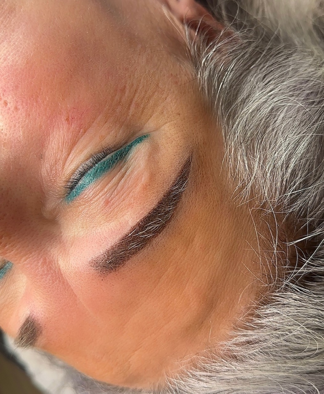 Zbliżenie na kobiecą twarz po zabiegu makijażu permanentnego brwi; widoczne świeżo wykonane, ciemne brwi i turkusowa kreska na powiece. Szare włosy w tle.