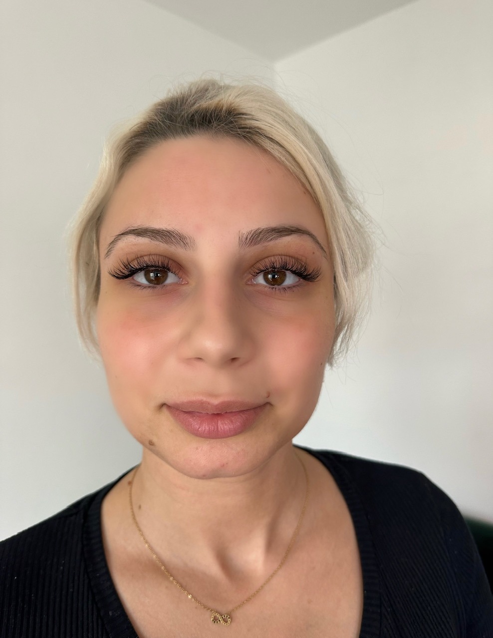 Portret kobiety z blond włosami i świeżo przedłużonymi rzęsami, widoczne brązowe oczy i złoty naszyjnik z symbolem nieskończoności.