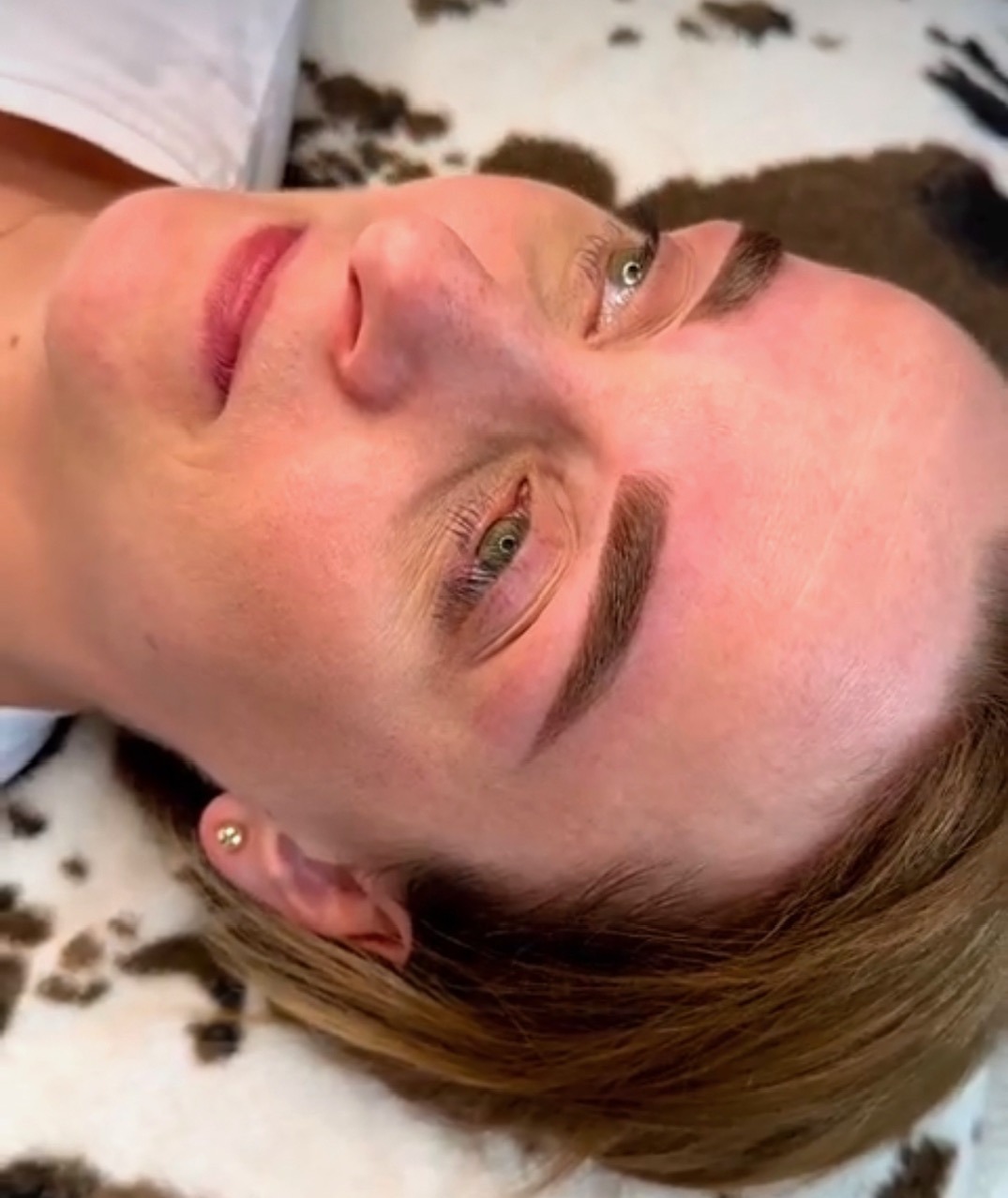 Kobieta po zabiegu microbladingu brwi, leżąca na wzorzystym podłożu. Widoczne świeżo wykonane, ciemne brwi i delikatny rumień na skórze.