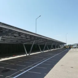 Zadaszony parking z panelami słonecznymi. Metalowa konstrukcja wsparta na słupach. W tle zieleń i błękitne niebo. Widoczni pracownicy w oddali.