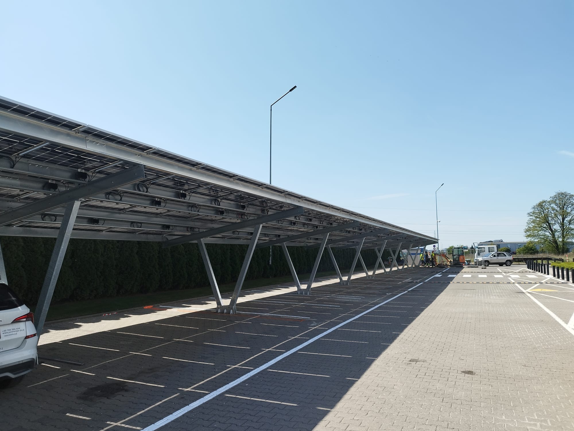 Zadaszony parking z panelami słonecznymi. Metalowa konstrukcja wsparta na słupach. W tle zieleń i błękitne niebo. Widoczni pracownicy w oddali.
