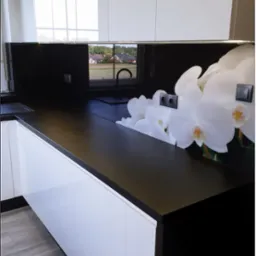 Nowoczesna kuchnia z białymi szafkami, czarnym blatem i kwiatowym motywem na ścianie. Widok na okno z zielonym krajobrazem w tle. Stylowe i funkcjonalne rozwiązanie.