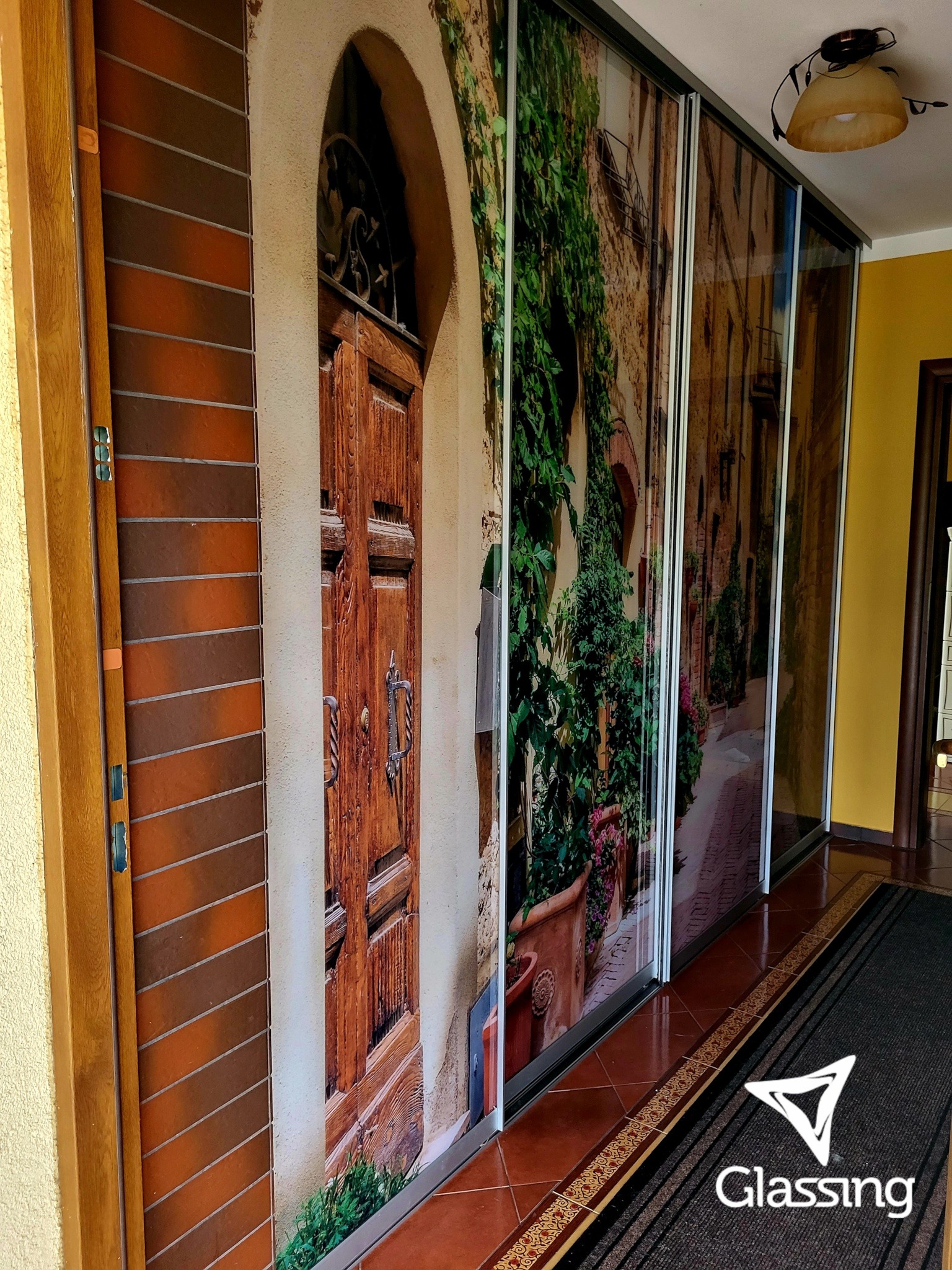 Szafa wnękowa z fototapetą imitującą włoską uliczkę i stare drzwi, wkomponowana w przedpokój z brązową podłogą i dywanikiem.