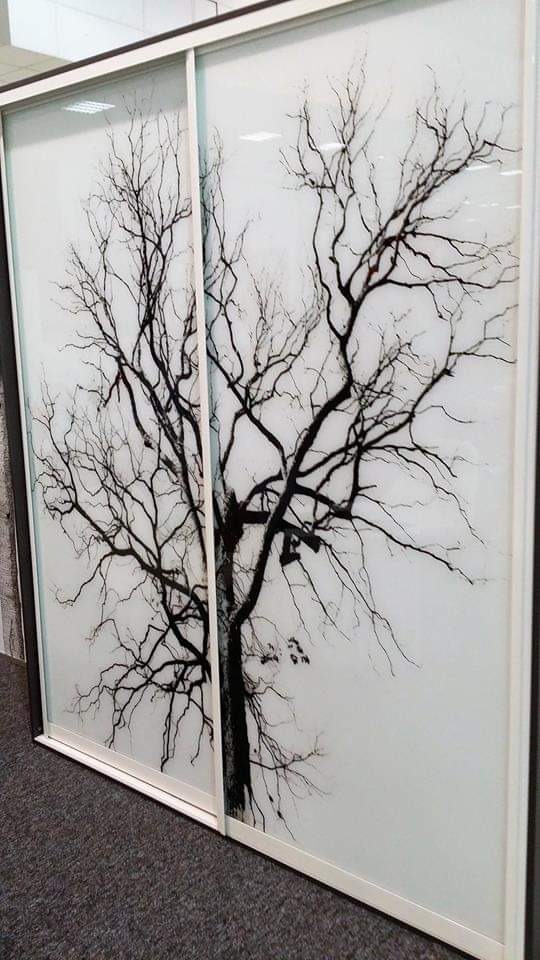 Biała szafa wnękowa z przesuwnymi drzwiami, na których widnieje czarny motyw drzewa bez liści, sięgający od podłogi do sufitu.