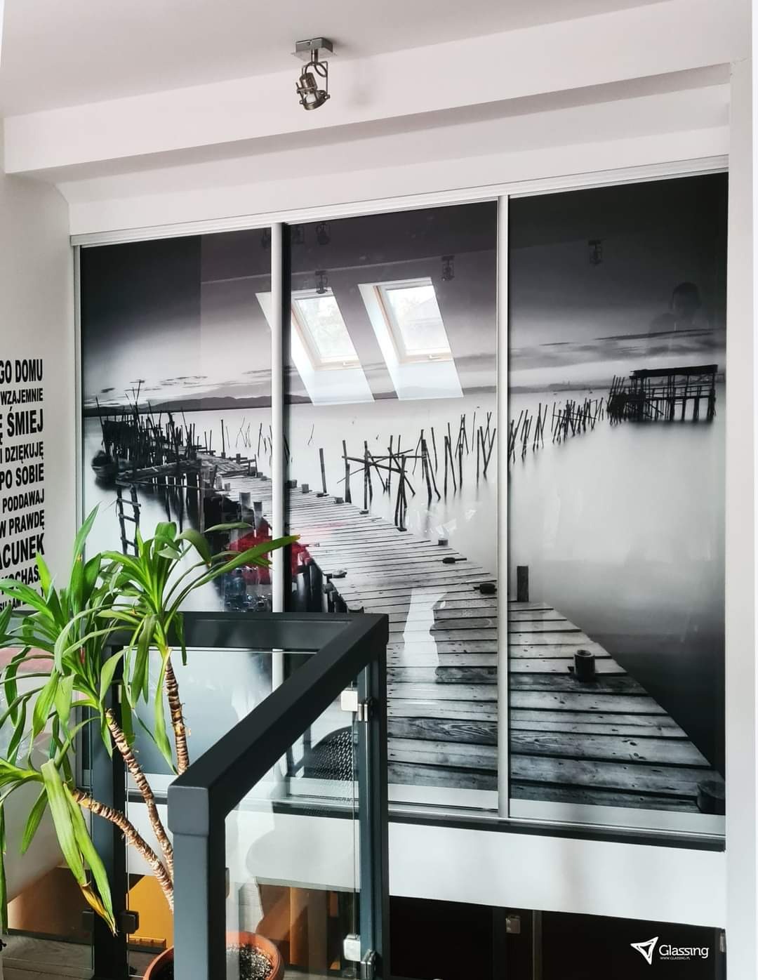 Czarno-biała grafika molo na szafie wnękowej z przesuwnymi drzwiami, widok z dołu, roślina doniczkowa z lewej strony, logo Glassing w prawym dolnym rogu.