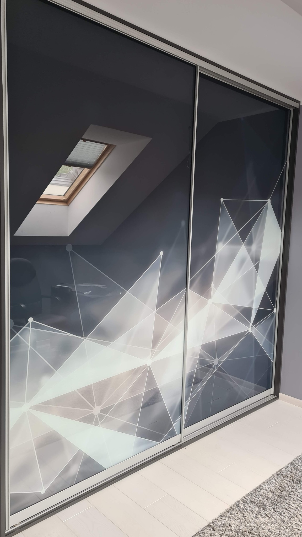 Dwudrzwiowa szafa wnękowa z przesuwnymi drzwiami, na których widnieje graficzny wzór białych linii na szarym tle. W tle okno dachowe.