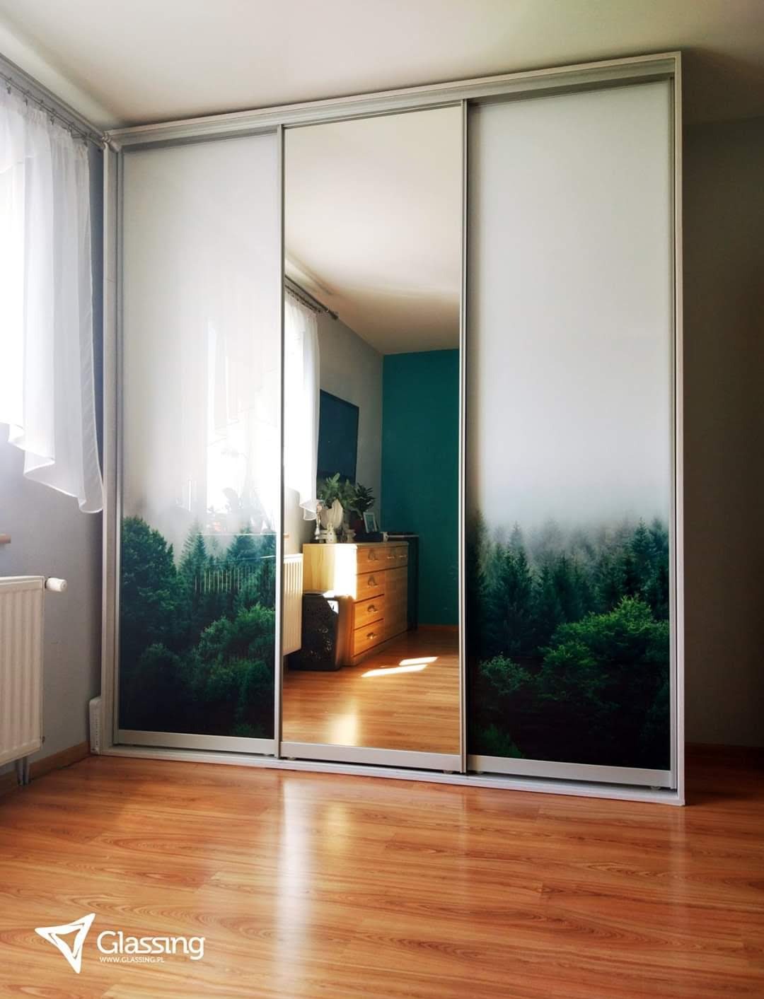 Szafa wnękowa z przesuwnymi drzwiami: lustro pośrodku i grafika lasu po bokach. Widoczne odbicie wnętrza pokoju i drewniana podłoga.