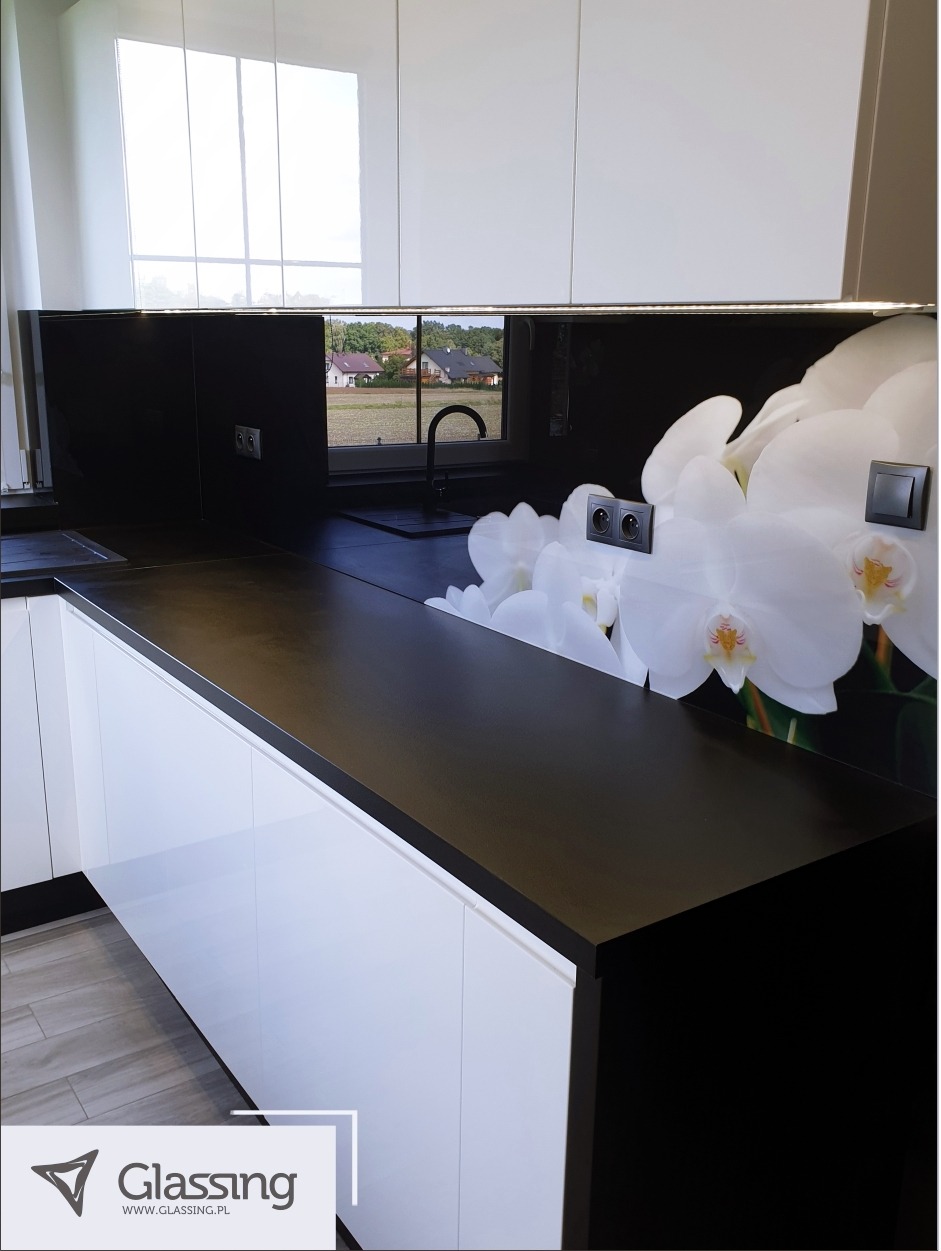 Nowoczesna kuchnia z białymi szafkami, czarnym blatem i kwiatowym motywem na ścianie. Widok na okno z zielonym krajobrazem w tle. Stylowe i funkcjonalne rozwiązanie.