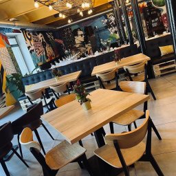 P.U.H. MARCIN SKRZYPICIEL - Ustawione stoły w restauracji z muralami, przygotowane na imprezę. Dekoracje kwiatowe na stołach, czarne sofy z palet. Industrialny wystrój wnętrza.