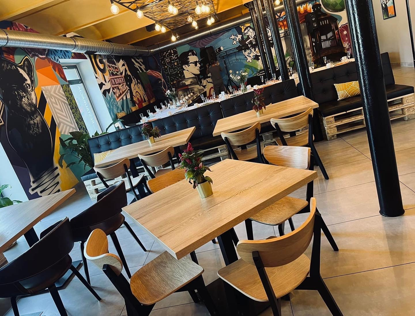 Ustawione stoły w restauracji z muralami, przygotowane na imprezę. Dekoracje kwiatowe na stołach, czarne sofy z palet. Industrialny wystrój wnętrza.