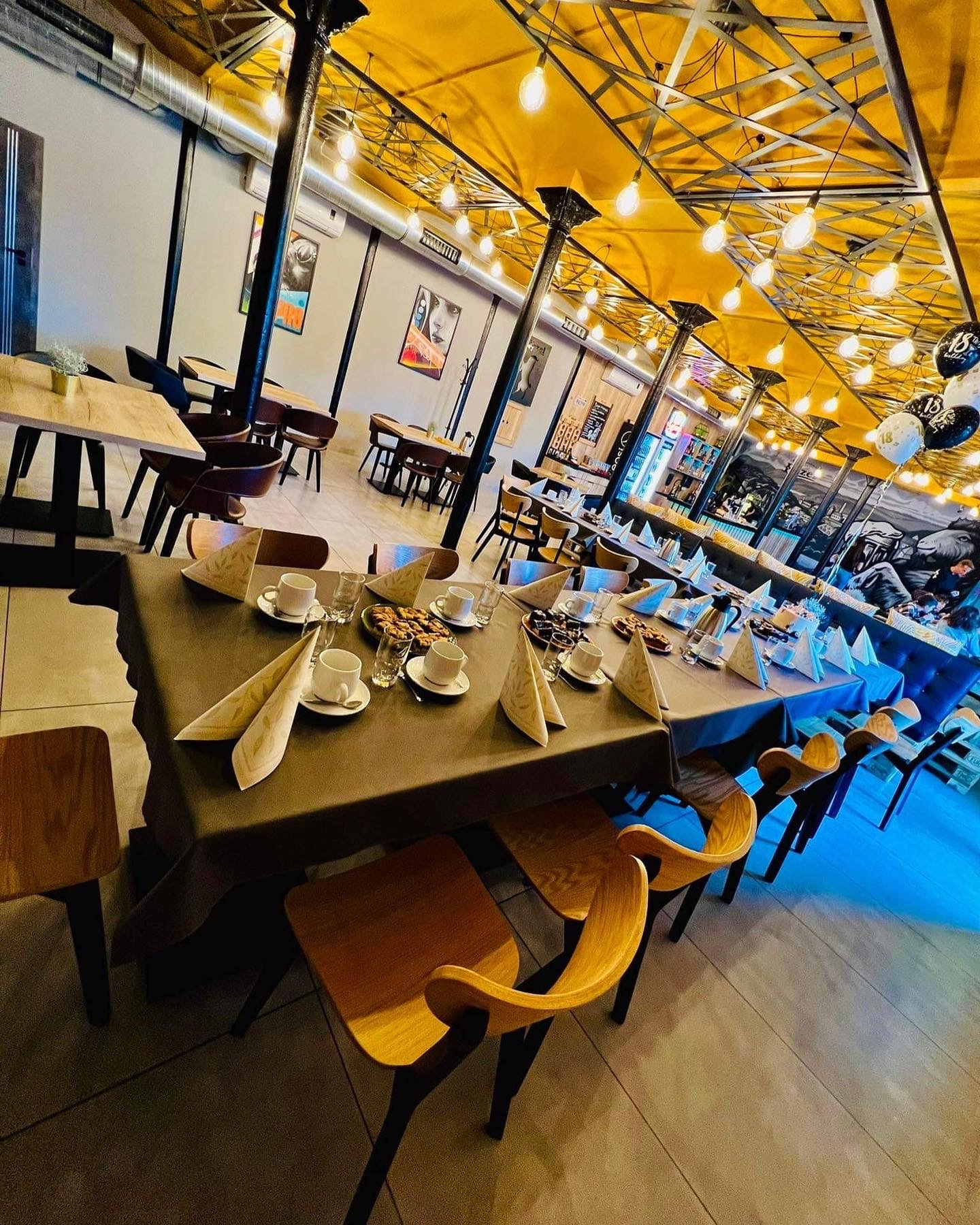 Uroczysty stół z nakryciami, kawą i ciastkami w jasnym wnętrzu z dekoracjami balonowymi, przygotowany na przyjęcie osiemnastkowe.