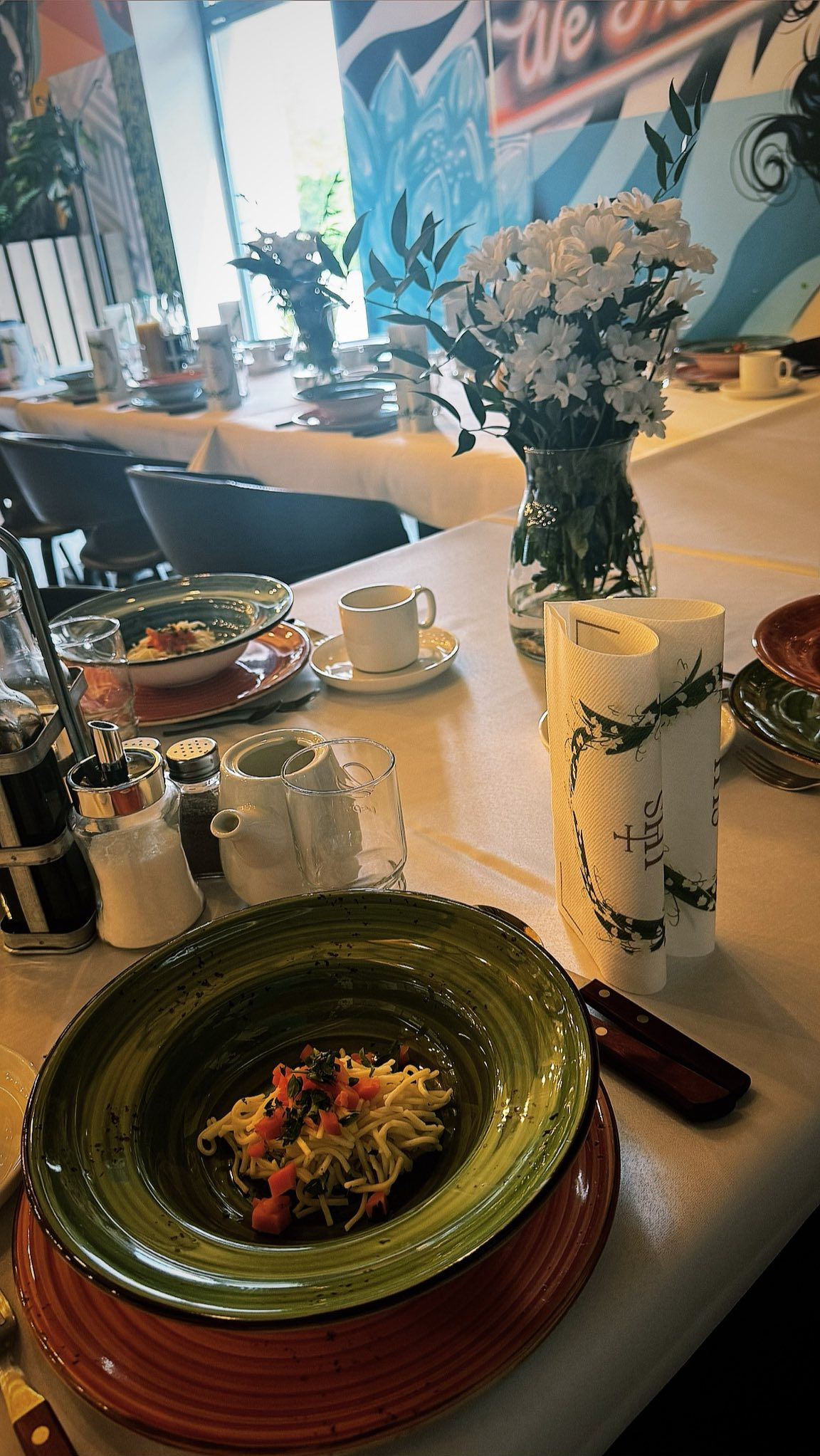 Uroczysty stół z zielonym talerzem spaghetti, kwiatami w wazonie, i eleganckimi serwetkami. Dekoracja stołu na imprezę z artystycznym muralem w tle.