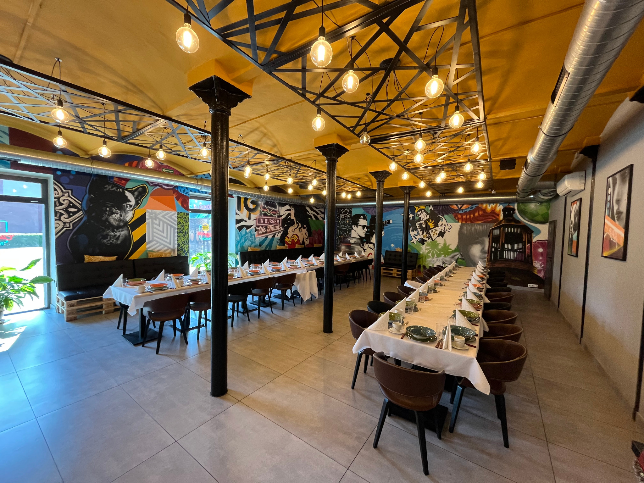 Sala bankietowa z długimi stołami nakrytymi do uroczystości, oświetlona dekoracyjnymi żarówkami, z muralami na ścianach i żółtym sufitem.