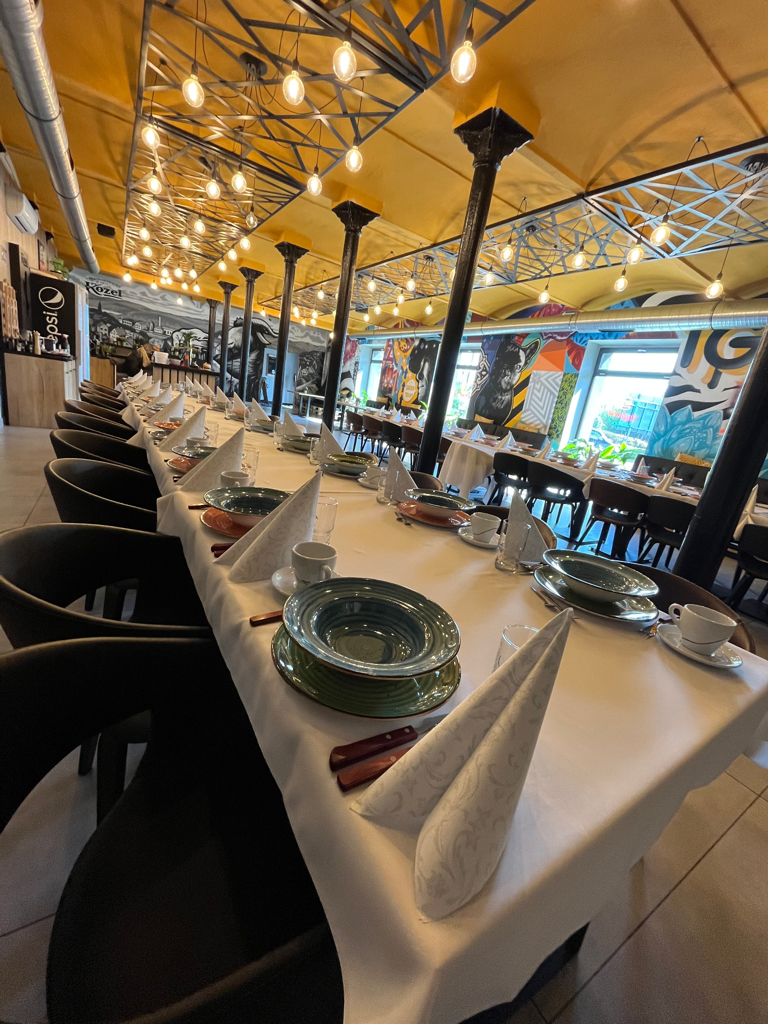 Uroczysty stół z zastawą i dekoracyjnymi serwetkami, przygotowany na przyjęcie w przestronnej sali z muralami i oświetleniem.