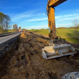 Żółta koparka CAT na placu budowy, obok drogi. Widoczne ślady pracy maszyny w ziemi, błękitne niebo z chmurami w tle. Prace ziemne w toku.