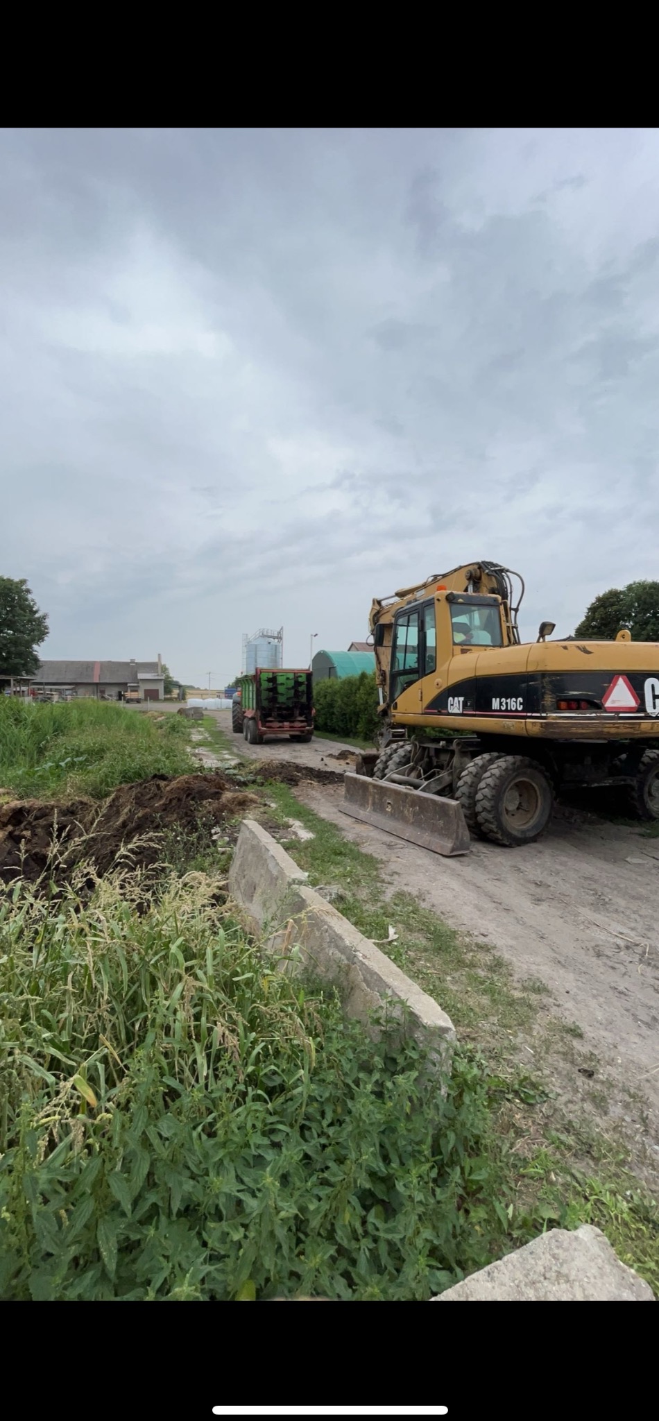 Żółta równiarka CAT M316C na polnej drodze, w tle zielony ciągnik z przyczepą. Wokół bujna roślinność. Pochmurne niebo.
