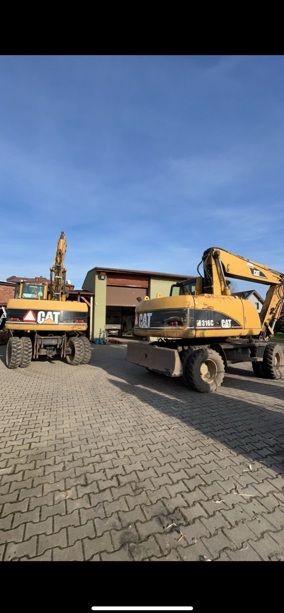 Dwie żółte koparki marki CAT na kostce brukowej przed budynkiem z brązową bramą garażową. Jedna z koparek to model M316C.