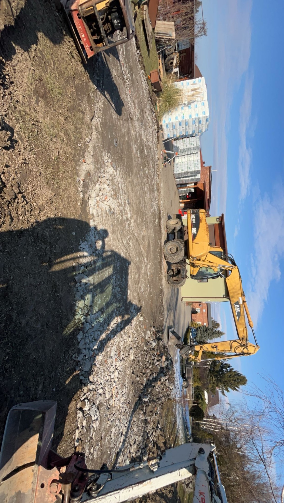 Żółta koparka na placu budowy, w tle budynki i niebo. Widoczny stos materiałów budowlanych, gruz i cień koparki. Ujęcie z góry, podkreślające skalę prac ziemnych.