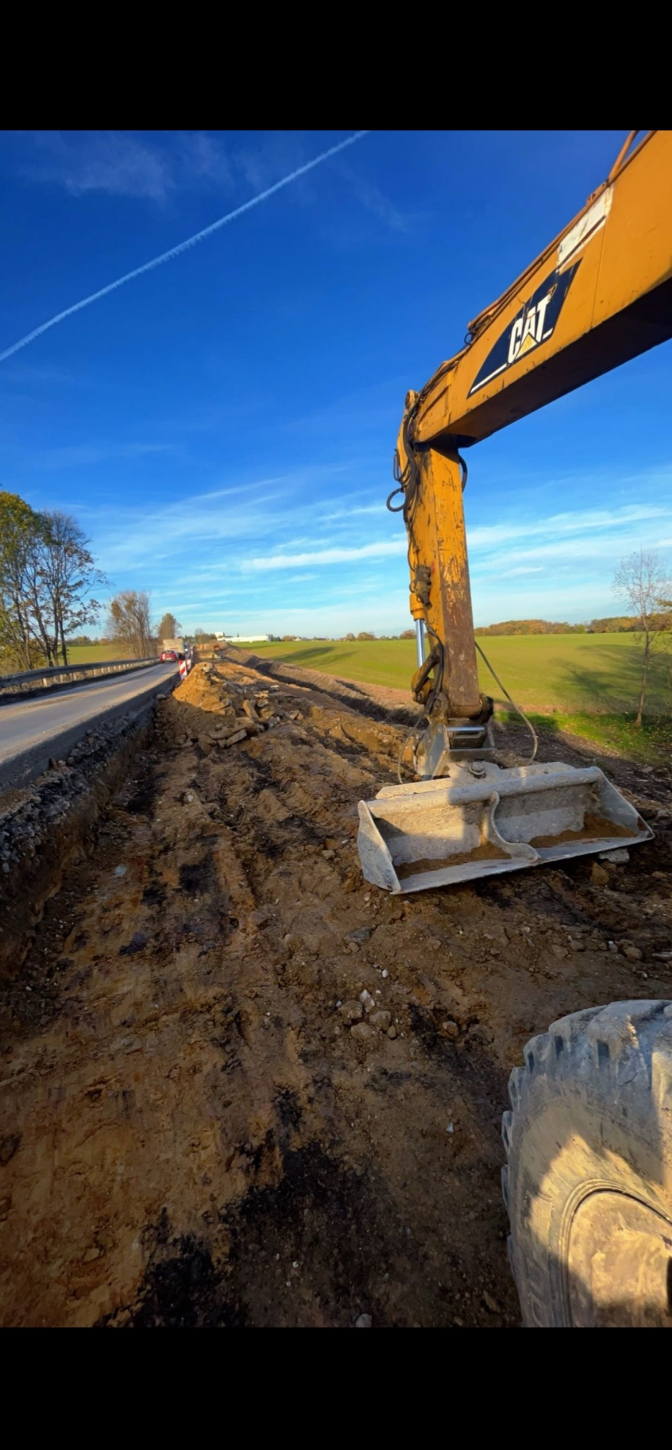 Żółta koparka CAT na placu budowy, obok drogi. Widoczne ślady pracy maszyny w ziemi, błękitne niebo z chmurami w tle. Prace ziemne w toku.