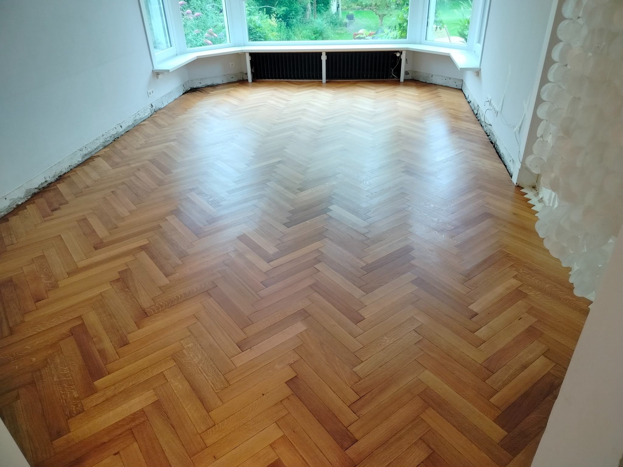 Parkiet ułożony w jodełkę w jasnym pokoju z oknem. Widoczne niedokończone ściany i listwy przypodłogowe, co sugeruje etap prac wykończeniowych.