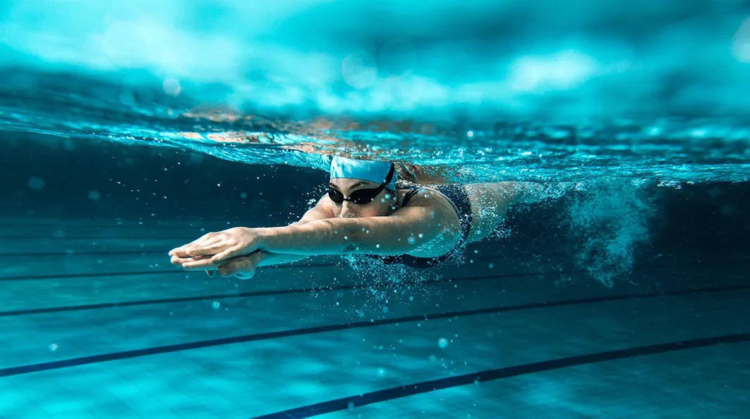 Podwodny kadr kobiety w czepku i okularach, płynącej stylem dowolnym w basenie z wyraźnie zaznaczonymi liniami torów, z bąbelkami powietrza wokół.
