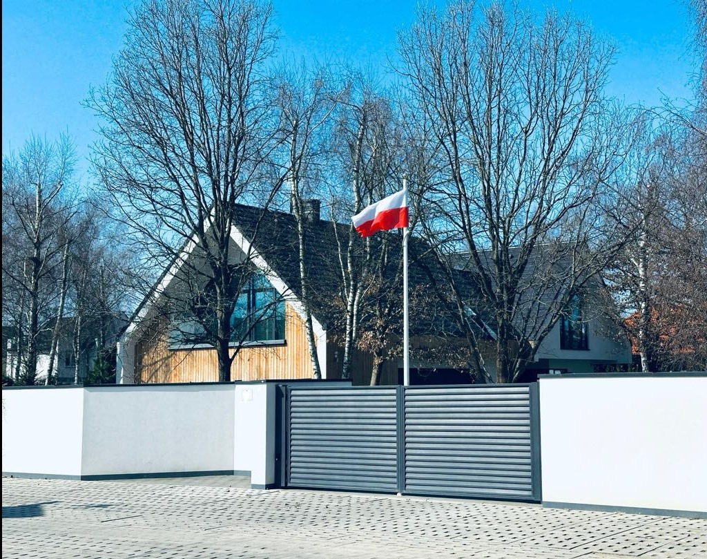 Nowoczesny dom z drewnianą elewacją, biało-szarym ogrodzeniem i bramą wjazdową. Na posesji powiewa polska flaga na tle błękitnego nieba i drzew bez liści.